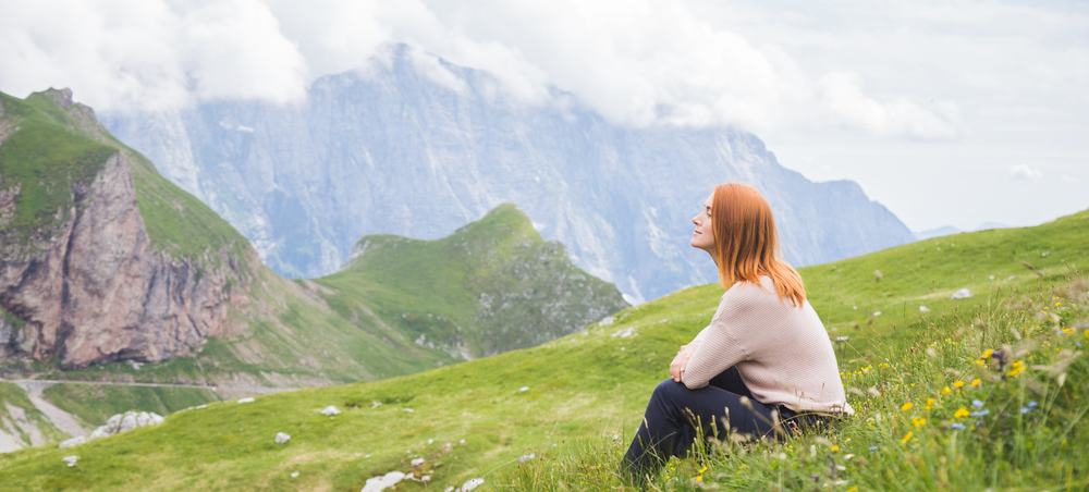 Portrait of a woman outdoors. Pretty young girl sitting in the grass outside, breathing fresh air and enjoying the sun. Mountain landscape with snowy peaks. Vacations in Europe national park.