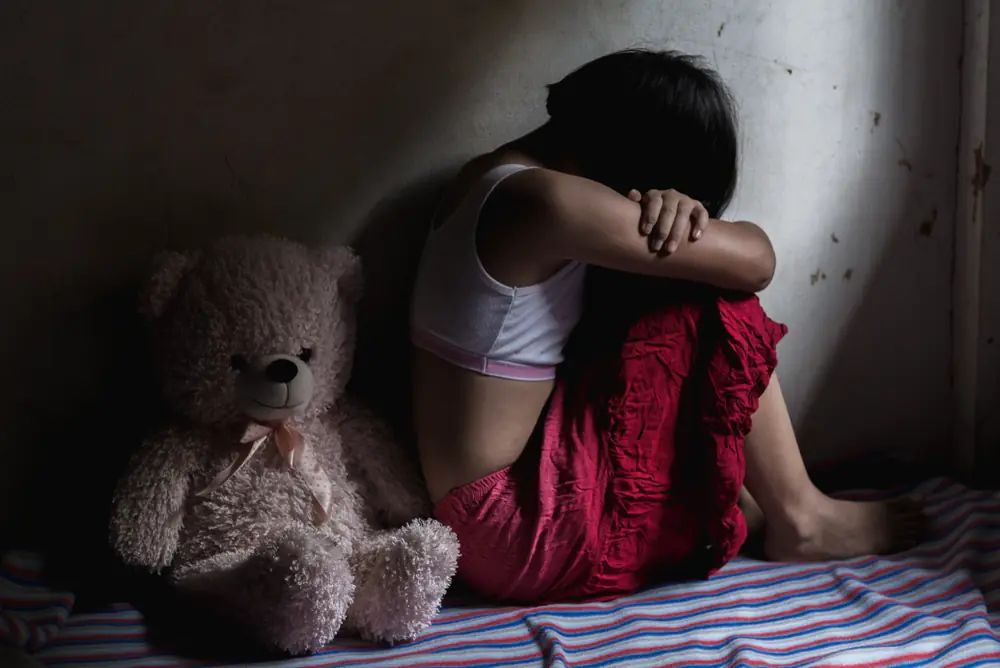 Person sitting on the floor with their head down on their knees beside a teddy bear